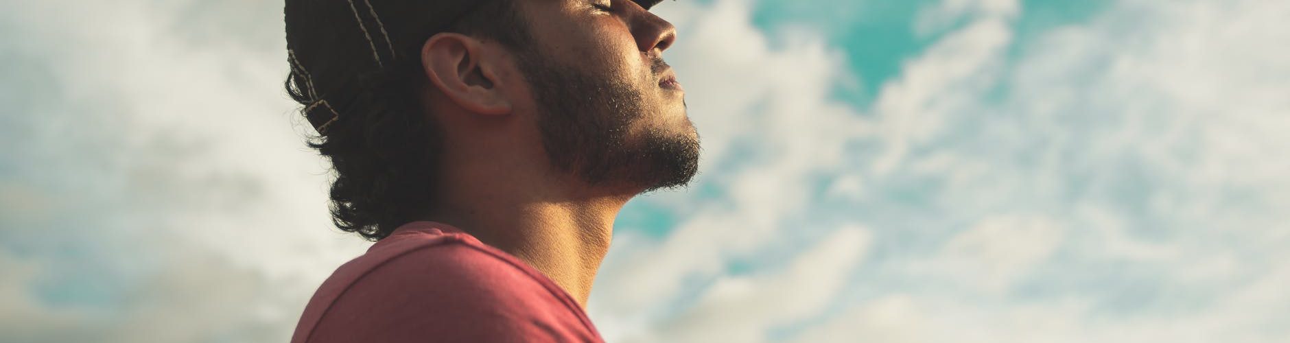 man wearing black cap with eyes closed under cloudy sky