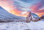 brown house surrounded by snow covered field near snow covered mountain under yellow blue and orange sunset