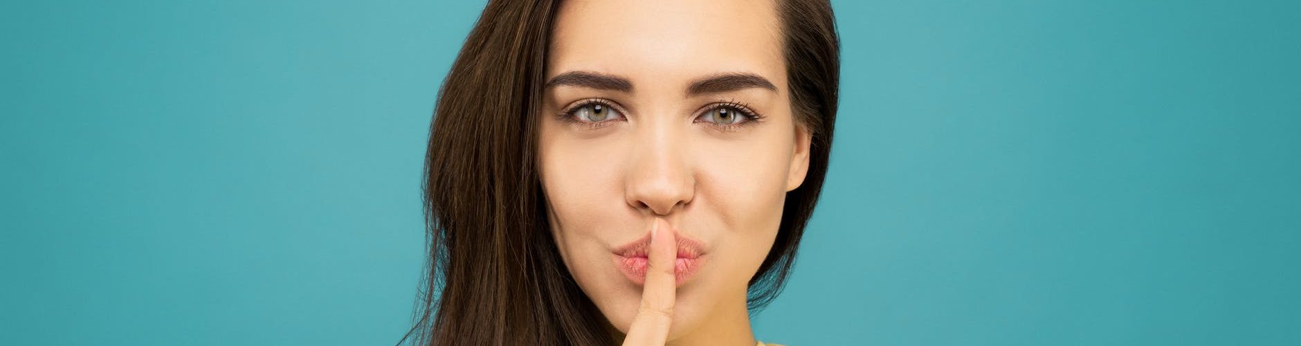 portrait photo of woman in yellow t shirt doing the shh sign while standing in front of blue background