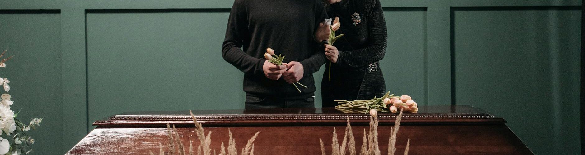 an elderly woman leaning on a man s shoulder near a wooden coffin
